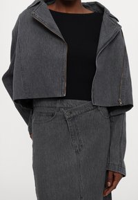 Giacca di jeans grigia corta con chiusura a zip e colletto ampio, abbinata a un top nero a coste e una gonna di jeans grigia a vita alta.