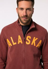 Giacca a zip color bordeaux con la scritta "ALASKA" in grassetto giallo sul petto, dotata di colletto alto e tasche frontali.