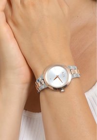 Hõbedane ja roosakuldne randme kell ümmarguse näo, metallist rihmaga ja minimalistliku disainiga. Omab peegeldavat tausta ja lihtsaid tunni näitajaid.