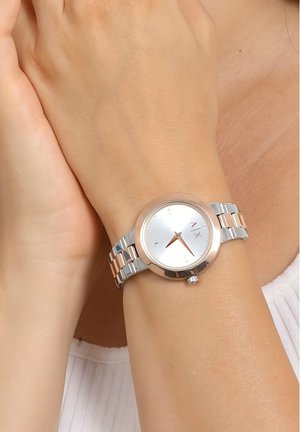 Hõbedane ja roosakuldne randme kell ümmarguse näo, metallist rihmaga ja minimalistliku disainiga. Omab peegeldavat tausta ja lihtsaid tunni näitajaid.