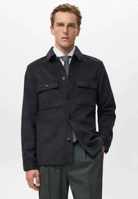 Dunkle, strukturierte Jacke mit zwei Brusttaschen, Knopfleiste und Kragen, getragen über einem hellen Hemd und einer Krawatte.