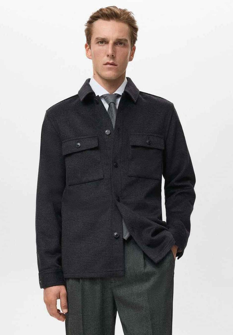 Dunkle, strukturierte Jacke mit zwei Brusttaschen, Knopfleiste und Kragen, getragen über einem hellen Hemd und einer Krawatte.