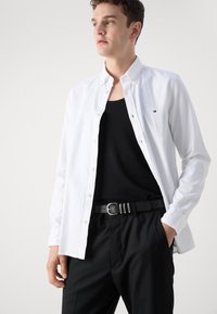 Camicia bianca con bottoni, in cotone, maniche lunghe, indossata sopra un top nero. Cintura nera con fibbia argentata, pantaloni neri. Design semplice.