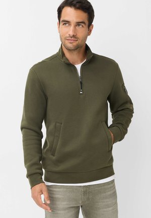 Mand iført olivengrønt kvart-zip pullover med hånden i lommen, kigger let til siden mod en ensfarvet lys baggrund.