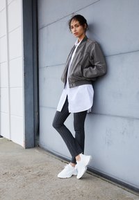 Grå bomberjakke over en hvid skjorte og sorte bukser med nålestriber, kombineret med hvide sneakers. Stoffet ser let ud; designet er afslappet.