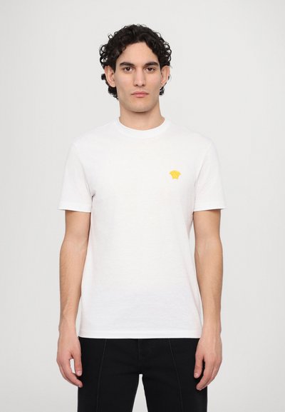 Witte katoenen t-shirt met korte mouwen, ronde halslijn en een klein geel geborduurd logo op de borst. Minimalistisch ontwerp.