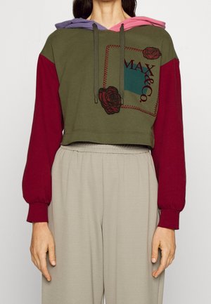 Vrouw draagt een cropped hoodie met kastanjebruine mouwen, olijfgroene voorkant met rozenpatches en de tekst "MAX&Co", gecombineerd met beige broek.