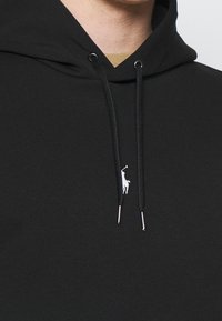 Czarna bluza z kapturem z przednią kieszenią typu kangur, regulowanymi ściągaczami zakończonymi srebrnymi końcówkami oraz białym haftowanym logo.