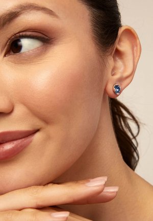 Pendientes de botón de gema azul en un bisel de tono plateado, que presentan una superficie suave y reflectante contra un fondo de piel clara.