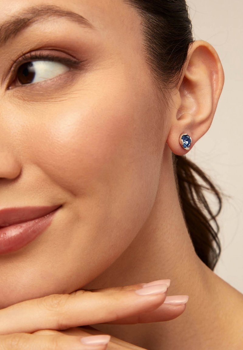 Pendientes de botón de gema azul en un bisel de tono plateado, que presentan una superficie suave y reflectante contra un fondo de piel clara.