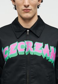 Νεαρό άτομο που φοράει μαύρο μπουφάν με φερμουάρ, με μεγάλο ροζ και πράσινο στυλιζαρισμένο κείμενο "ICECREAM" στο στήθος.