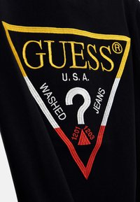Svart tyg med djärvt gult, vitt och rött broderat triangellogo. Innehåller texten "GUESS U.S.A. WASHED JEANS" och ett frågetecken.