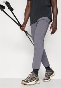 Gray outdoor pants, black and beige trail shoes with a textured sole, and trekking poles held in one hand, on a plain background.