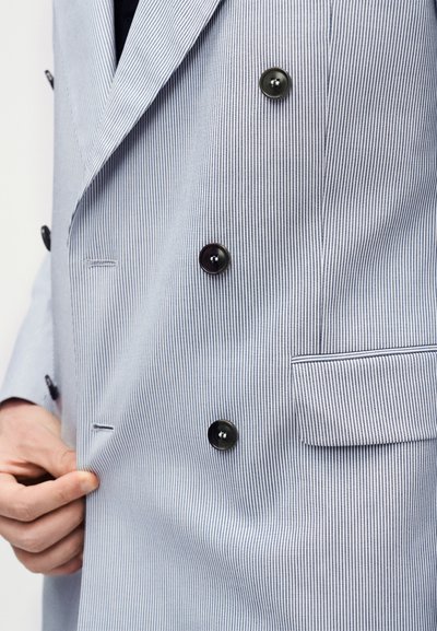 Άτομο που διορθώνει ένα ανοιχτό μπλε σακάκι με λεπτές ρίγες, διπλής ραφής, με μαύρα κουμπιά και τσέπη με καπάκι στο πλάι.