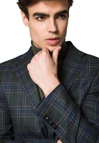 Kratkowany wełniany żakiet w ciemnozielonym kolorze z niebieskimi i brązowymi akcentami. Posiada dwa guziki rękawów oraz klapy. Model pozuje w zamyśleniu.