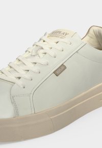 Gros plan d'une basket de couleur crème de la marque Replay avec des lacets et une semelle en caoutchouc beige sur fond blanc.