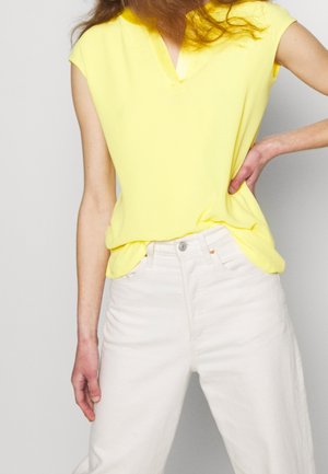 Femme portant un haut jaune à manches courtes rentré dans un pantalon blanc taille haute, avec une main sur la hanche et l'autre détendue.
