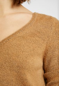 Pull en maille marron avec un décolleté en V. Texture douce avec une finition légèrement duveteuse. Présente un tissage lisse et uniforme.