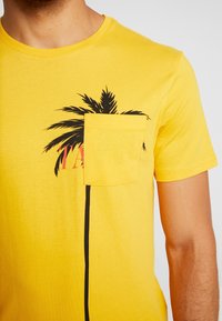 Žluté tričko s krátkým rukávem se vzorem palmy, náprsní kapsou a černým pruhovaným detailem, který probíhá vertikálně podél postranního švu.
