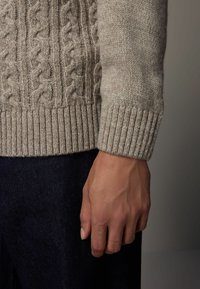 Beiger Cable-Knit-Pullover mit gerippten Bündchen, weicher Textur und lockerer Passform, kombiniert mit dunkelblauen Jeans. Nahaufnahme von Arm und Hand.