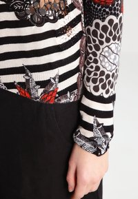 Hand vilar på en svart kjol, iförd en långärmad topp med svart- och vitrandigt mönster samt röda och bruna blommor och spetsmönster.