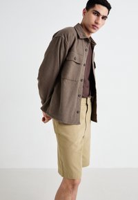 Brown textured shirt jacket with button closure, two chest pockets, paired with beige shorts. Simple design, casual fit.
