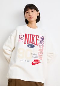 Balts, pārāk liels, sporta krekls ar sarkaniem un krēmkrāsas Nike grafiskajiem elementiem, ieskaitot "1972", "90" un logotipus. Mīksta auduma, ar ribotām apkaklītes un piedurkņu malām.