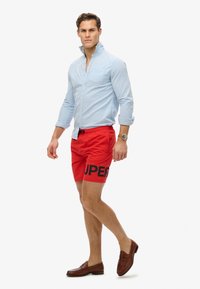 Mand iført lys blå skjorte med knapper, røde shorts med teksten "SUPER", brune loafers og et armbåndsur mod en ensfarvet hvid baggrund.