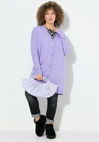 Cardigan long violet avec un devant à boutons, des poignets côtelés, associé à un jean noir et des bottes noires, tenant un sac violet clair.