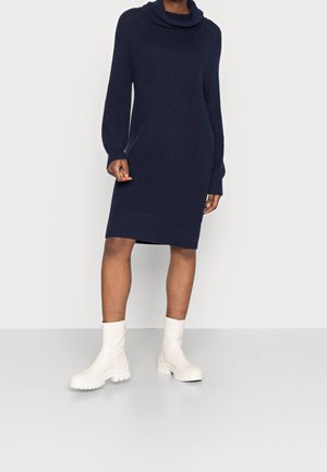 Person wearing a navy blue knitted long-sleeve sweater dress with a cowl neck and white chunky ankle boots, standing on a plain floor.