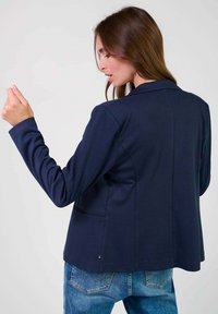 Blazer blu navy con un design su misura, caratterizzato da maniche lunghe, colletto con punta e tasche laterali, abbinato a jeans in denim blue chiaro.