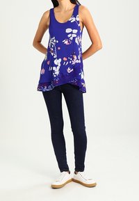 Fioletowy top bez rękawów z kwiatowym wzorem, luźny fason, warstwowy dół, zestawiony z ciemnymi dżinsami i białymi trampkami.