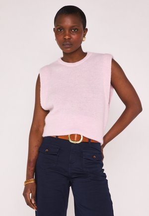 Frau mit kurzen Haaren, die einen ärmellosen hellrosa Pullover, marineblaue Hosen mit Vordertaschen, einen braunen Gürtel, goldene Ohrringe und Armbänder trägt.