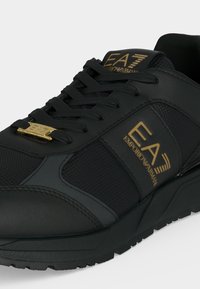 Μαύρο αθλητικό παπούτσι EA7 Emporio Armani με χρυσό λογότυπο στην πλαϊνή επιφάνεια και τη γλώσσα, μαύρα κορδόνια και υφασμένο πάτο.