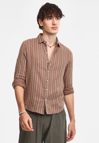 Camicia a maniche lunghe marrone a righe con chiusura a bottoni, tessuto morbido, maniche arrotolate e dettagli bianchi. Indossata con pantaloni verdi a vestibilità comoda.