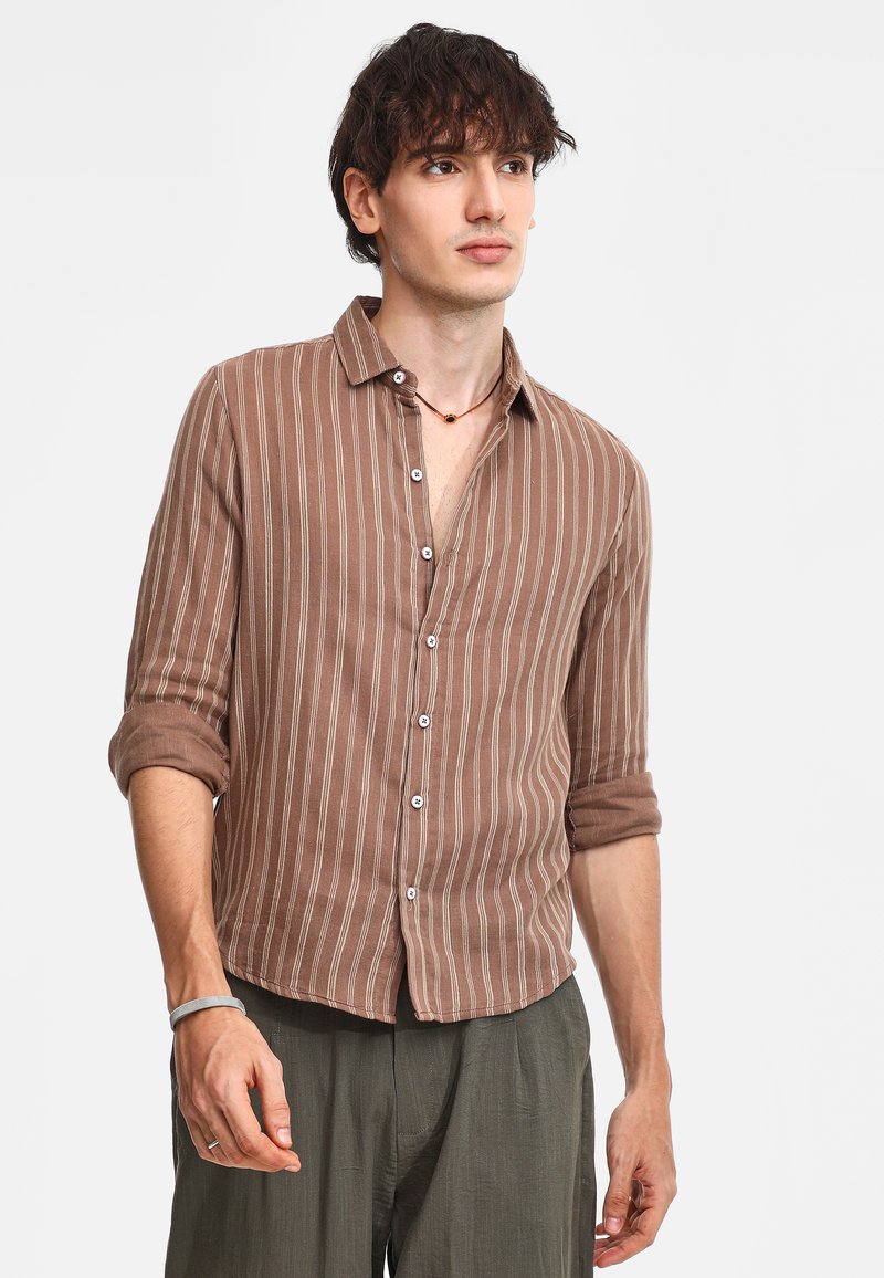 Camicia a maniche lunghe marrone a righe con chiusura a bottoni, tessuto morbido, maniche arrotolate e dettagli bianchi. Indossata con pantaloni verdi a vestibilità comoda.