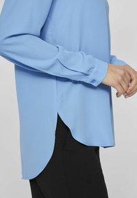 Lyseblå bluse med lange ermer og rund falm, med knaplukninger ved håndleddene, lavet af glat stof, kombinert med sorte bukser.