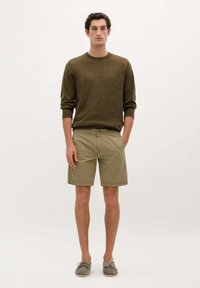 Pulover tricotat de culoare verde măslin, cu un finisaj texturat, asociat cu pantaloni scurți bej, până la genunchi. Modelul poartă pantofi de bărbat gri de tip boat, cu mâinile în buzunare.