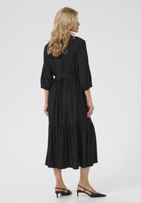 Robe noire à manches longues avec taille froncée, jupe à volants et légère brillance. Portée avec des chaussures à talons, noires et à lanières.