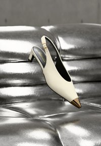 Λευκό γόνατο με λουράκι στο πίσω μέρος και χρυσή μύτη, τοποθετημένο σε λαμπερή ασημί επιφάνεια από δερμάτινο πλεκτό ύφασμα.