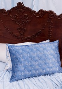 Blaues Satin-Kissen mit gesteppter Schleifenmusterung, begleitet von einem weißen Kissen. Vor einem kunstvoll verzierten, dunklen Holz-Kopfteil mit floralen Schnitzereien.