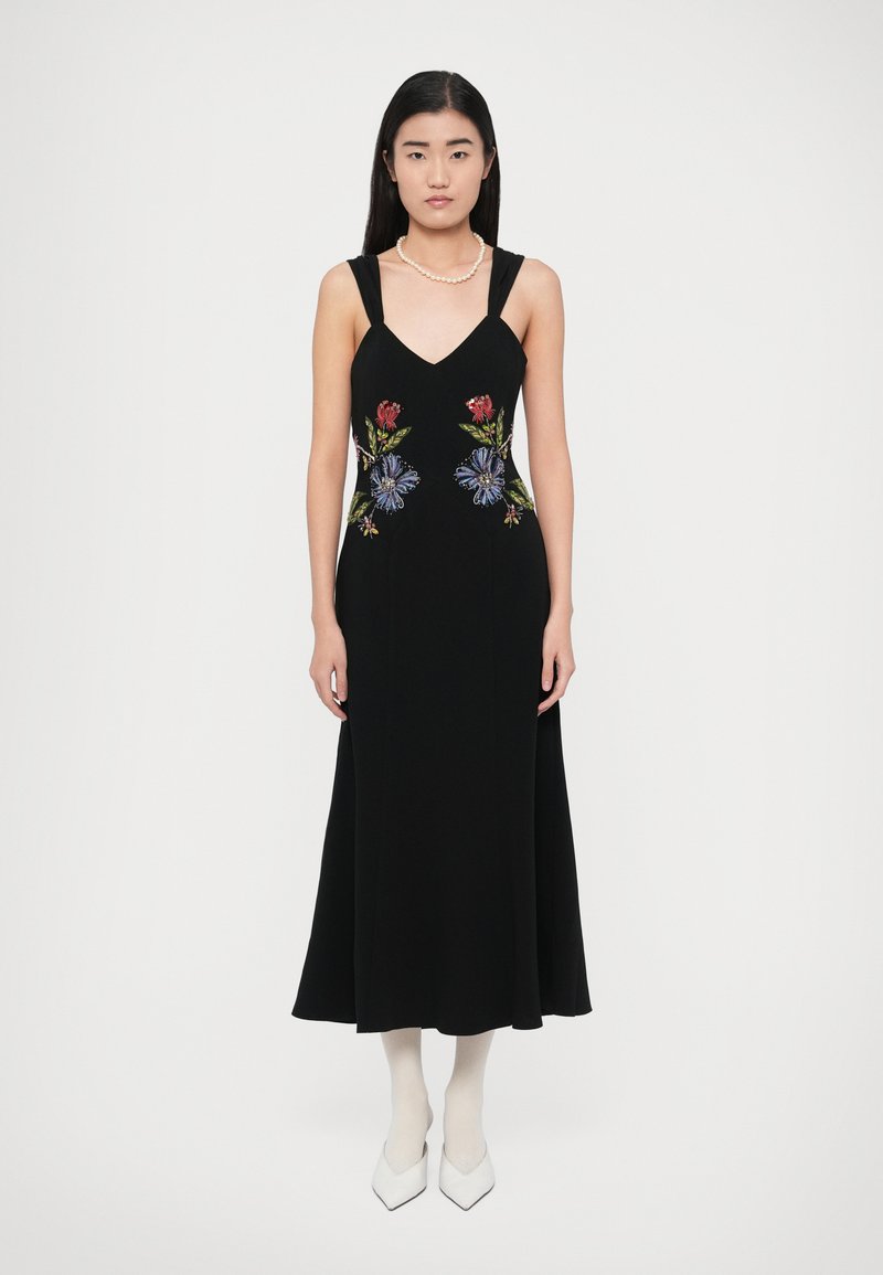 Juoda rankovių neturinti suknelė su raudonų, mėlynų ir žalių gėlių siuvinėjimais. Laidi silueto su V formos iškirpte; derinama su baltais smailiais aukštakulniais.