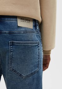 Jean en denim bleu avec une texture lisse, comportant une poche arrière et une étiquette beige portant l'inscription "PULL&BEAR DENIM DEPT."