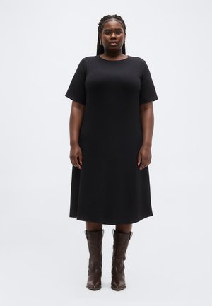 Kvinne med flettet hår iført en svart kjole til knærne og mørkebrune skinnstøvler som rekker til midt på leggen, stående med armene avslappet ned langs sidene.