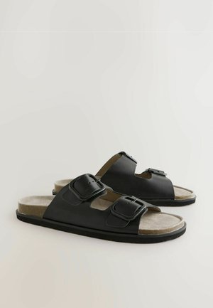 Zwarte slide-sandalen met twee verstelbare gespen, zachte bruine binnenvoering en geribbelde rubberen zool. Gladleren bovenwerk met een open teen ontwerp.