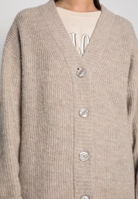 Cárdigan beige de punto con cuello en V, textura acanalada y cinco grandes botones de color claro en la parte delantera. Material suave y grueso.