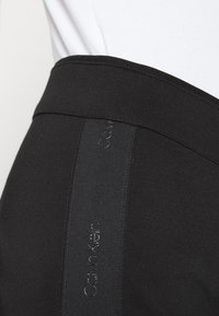 Svarta högmidjade leggings med en slät yta, som har en subtil tryckdetalj längs sidan och ett brett elastiskt midjeband.