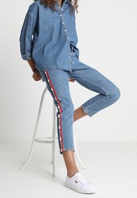 Jeansskjorta med långa ärmar och knäppning, i kombination med blå jeans med röda och svarta sidostreck, kompletterad med vita sneakers.