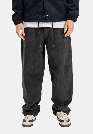 Mann trägt lockere dunkelgraue Cordhosen, weiße Sneakers mit schwarzen Streifen, schwarze Jacke und weißes Hemd vor einfachem Hintergrund.