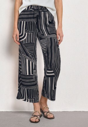 Kvinde iført sorte bukser med vide ben med hvide geometriske striber og sorte sandaler med gyldne detaljer, stående op ad en hvid væg.